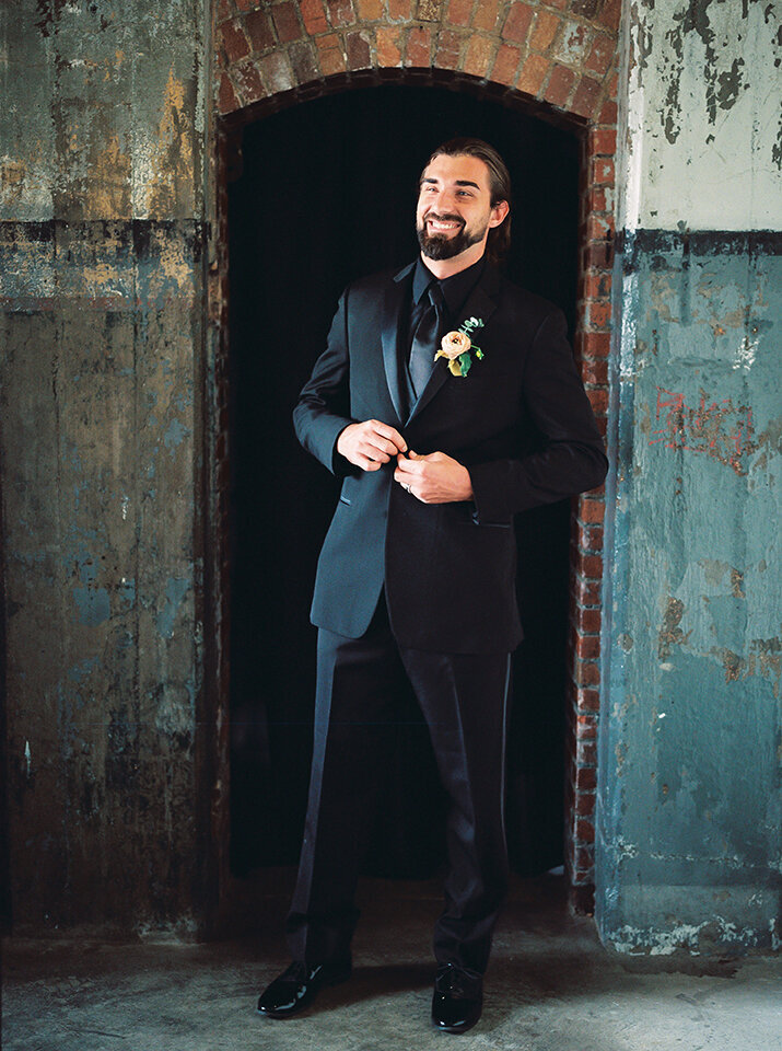 A groom wearing a black tuxedo smiles in front of a doorway along a weathered brick and wooden wall.