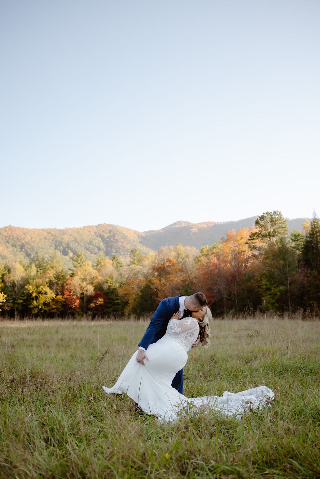 gatlinburg photographer captures groom dipping his bride backwards and kissing her passionately in a field in Cades Cove with the Smoky Mountains in the distance  