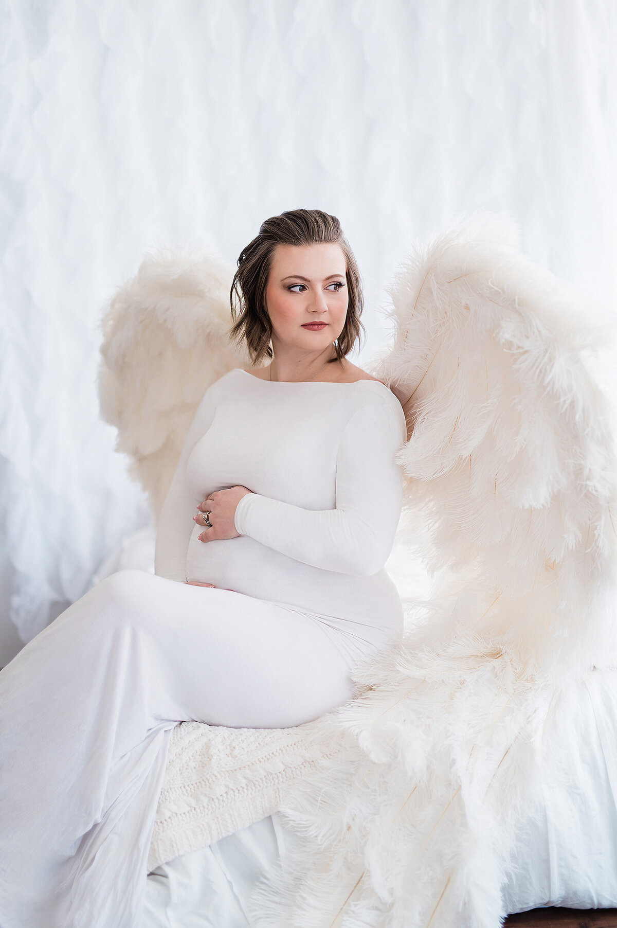 Pregnant woman sitting on a bed wearing a long white, boatneck maternity gown and angel wings made from white feathers.