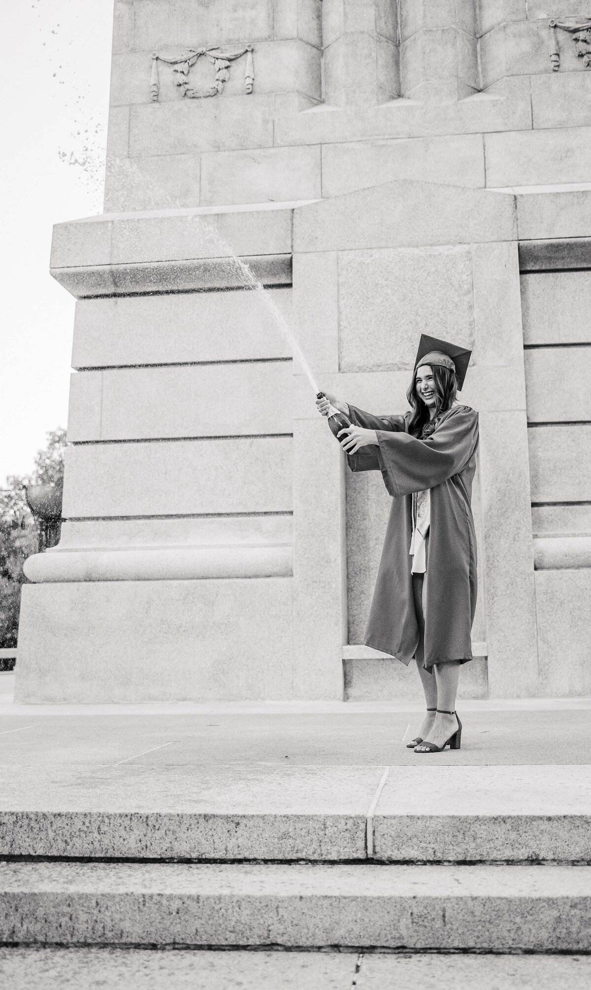 A graduate, captured by a senior portrait photographer from Fayetteville NC, stands jubilantly on the steps in cap and gown, spraying champagne. The black-and-white image beautifully frames a large stone structure in the background.