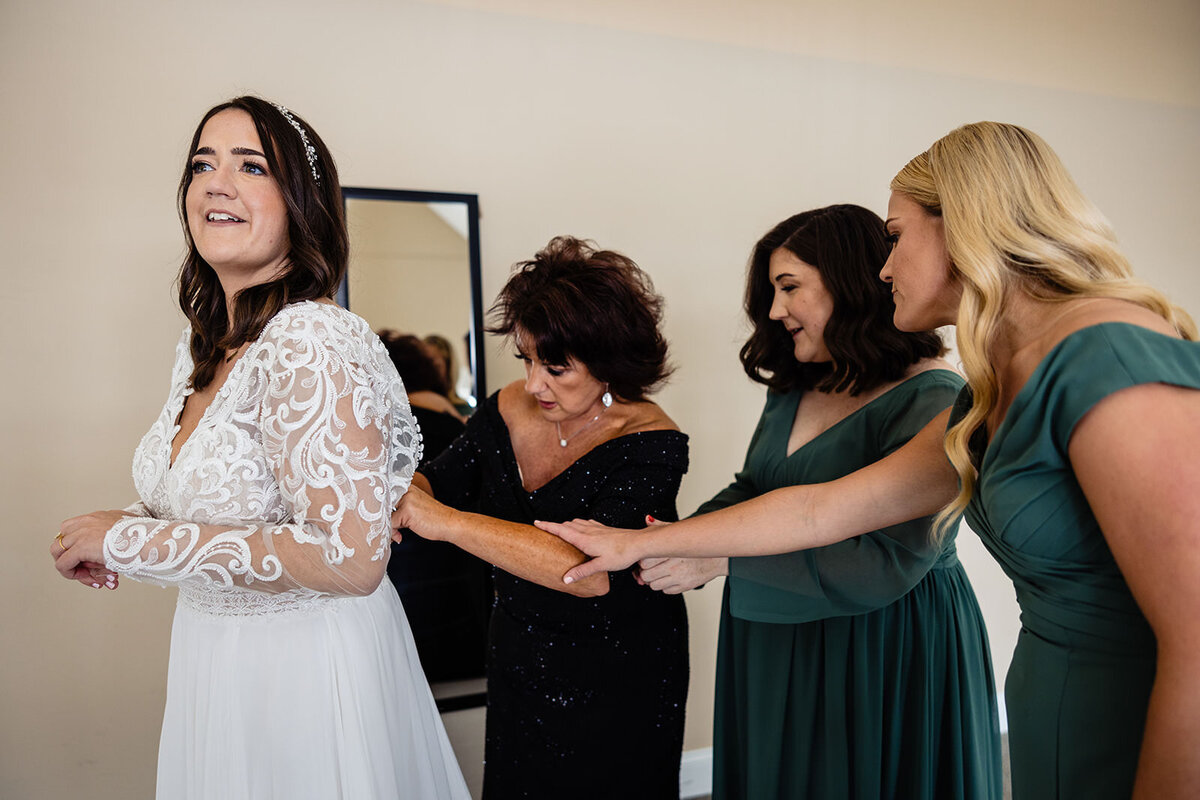 A bride in a lace wedding dress smiling while being helped into her gown by three women in a bright room.