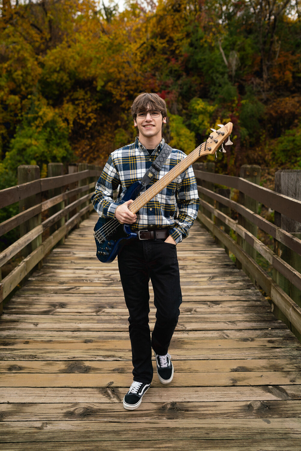 Senior boy walks across a bridge holding a bass guitar.