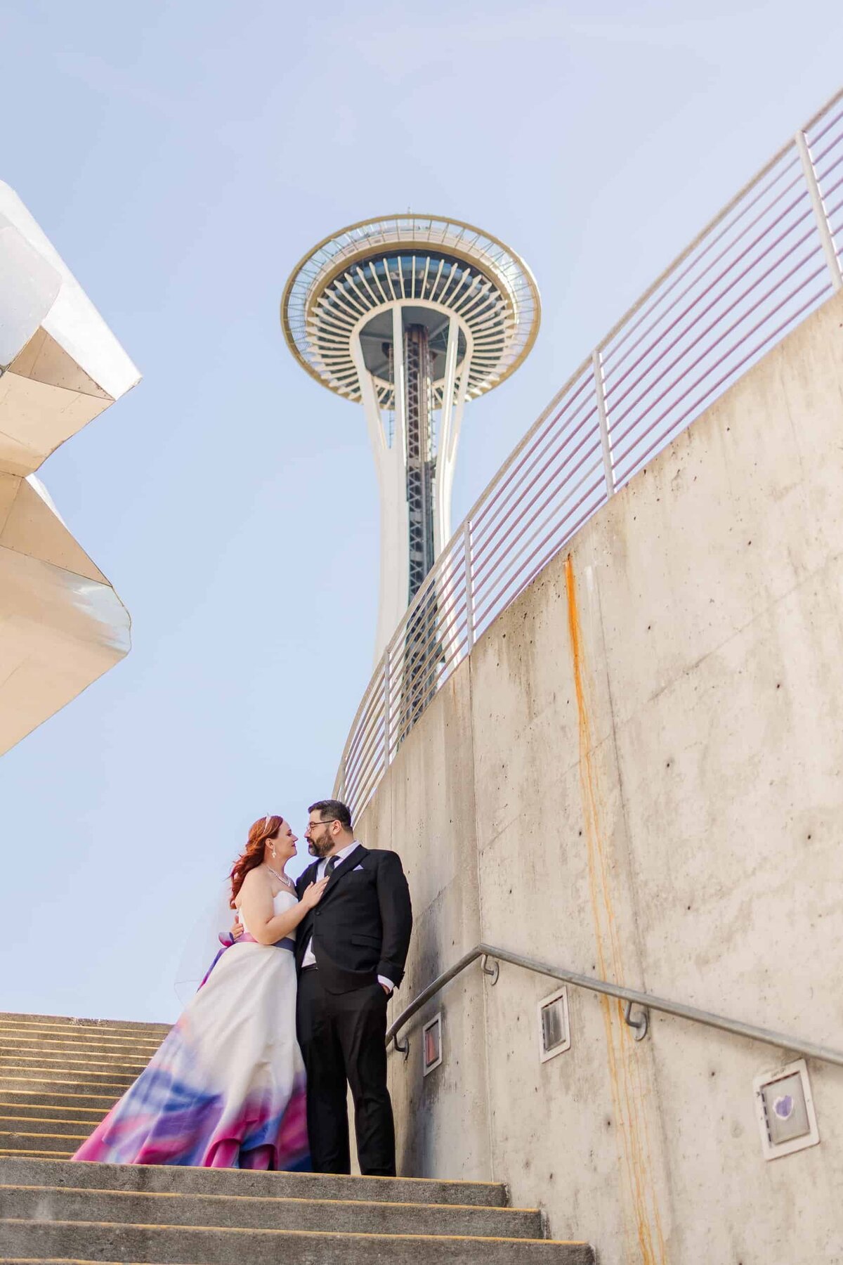 Couple in warm embrace, with bride, wearing non-traditional wedding dress with many colors, in front of Seattle's space needle