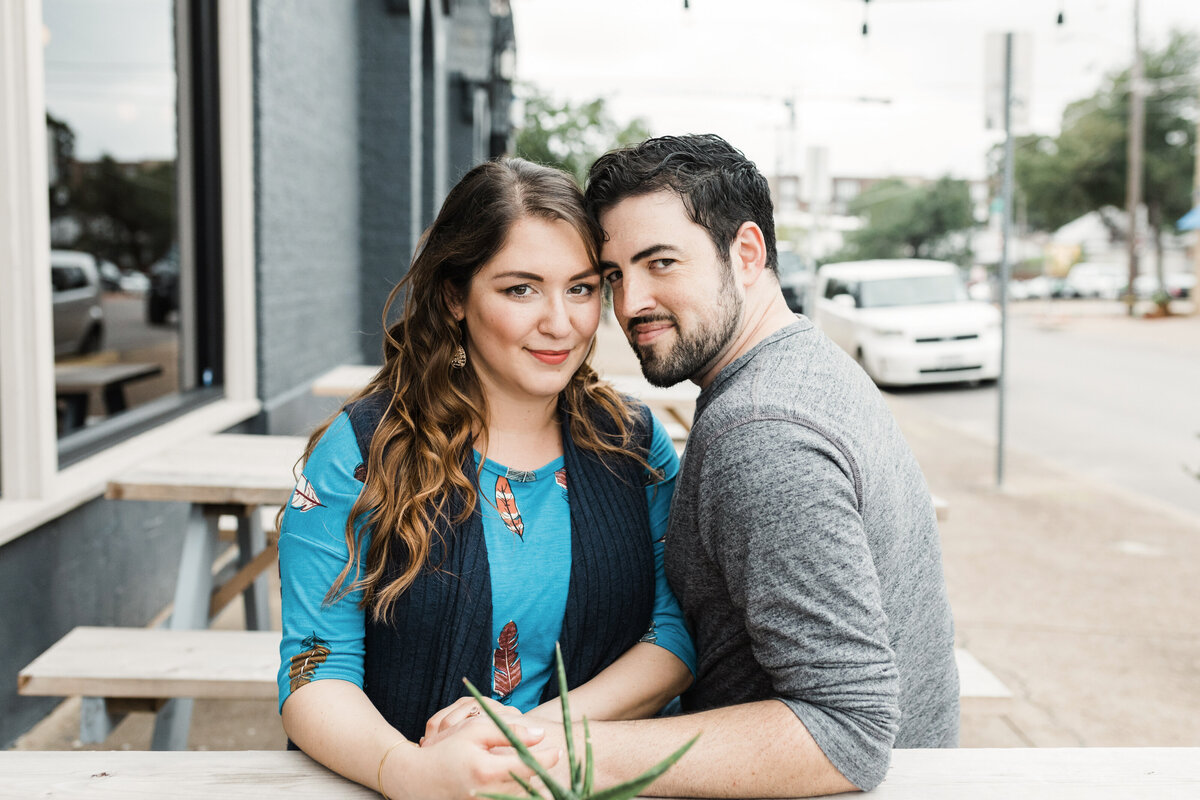A couple looking coyly at the camera while sitting at a picnic table during their engagement session in the Bishop Arts District in Dallas, Texas. The woman on the left is wearing a blue shirt decorated with leaves and a dark blue vest. The man on the right is wearing a grey shirt.