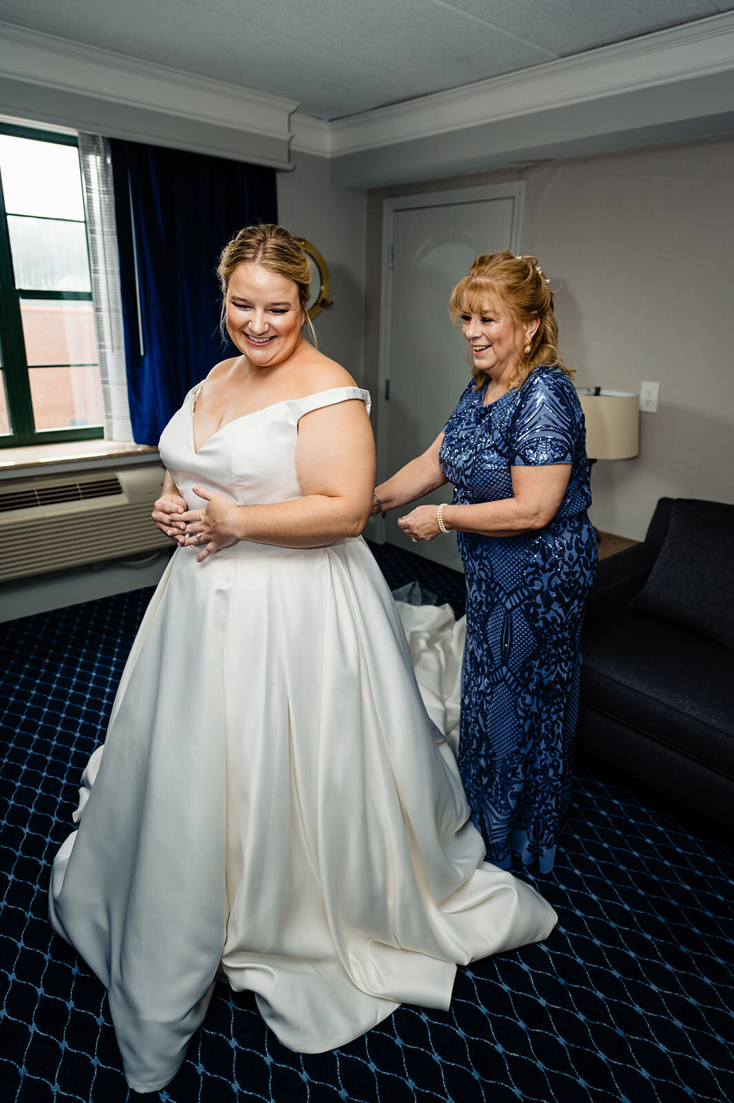 A smiling woman in a wedding gown being assisted by another woman in a blue lace dress