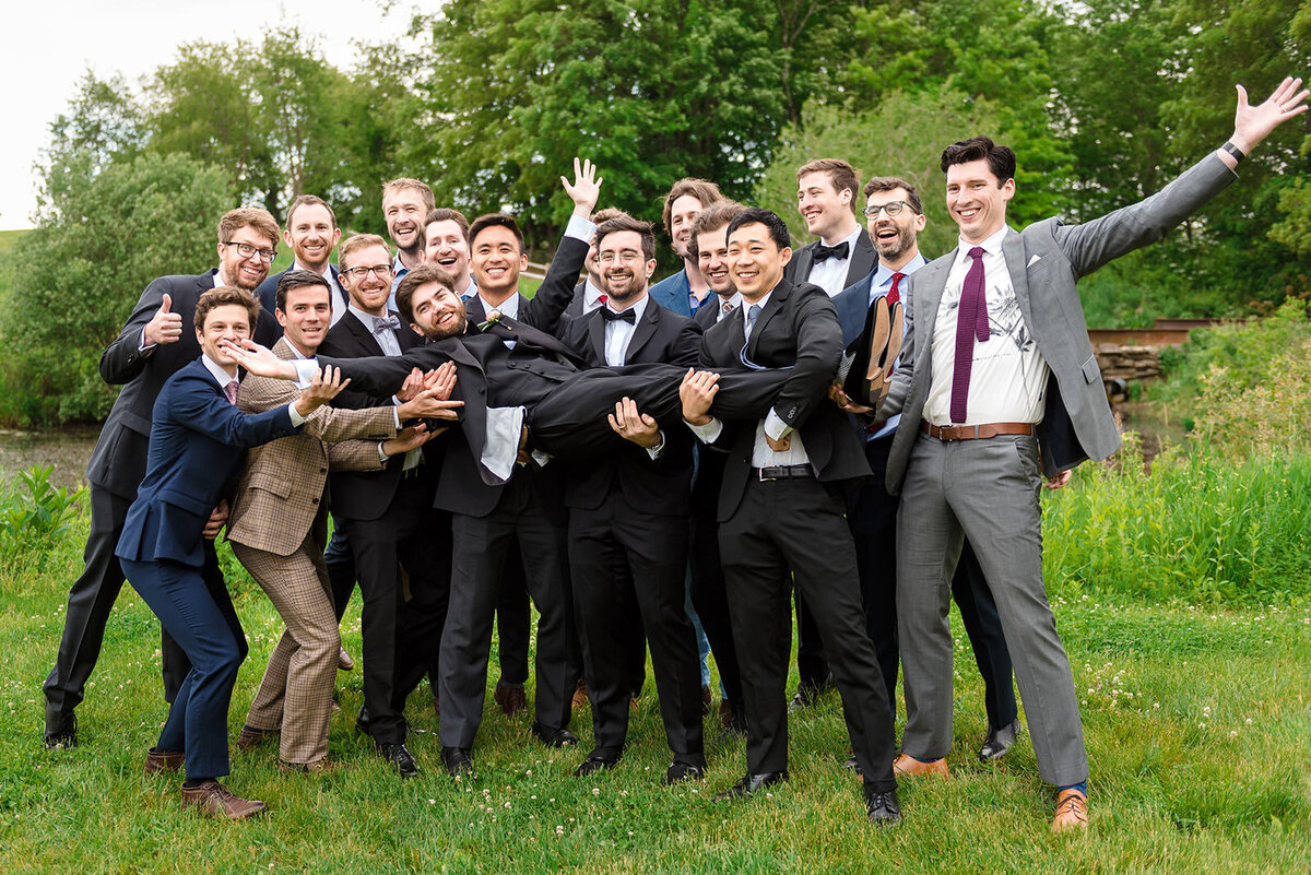 A group of groomsmen lifting the groom in a fun and playful wedding photo, outdoors on a grassy field.