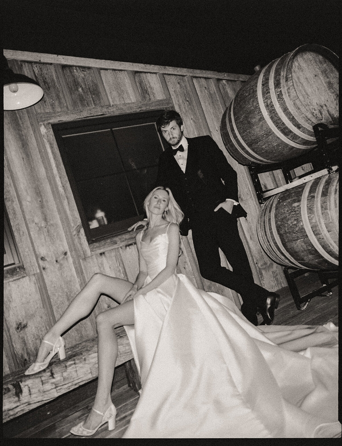 A stylish couple in formal attire, with a man standing and a woman sitting on barrels, posing dramatically in a rustic wooden room at an Iowa wedding.