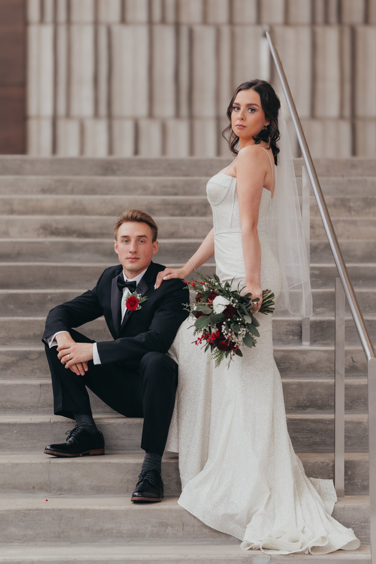 A bride and groom elegantly dressed, sitting on steps at a park farm winery wedding, with the bride holding a bouquet and looking at the camera.