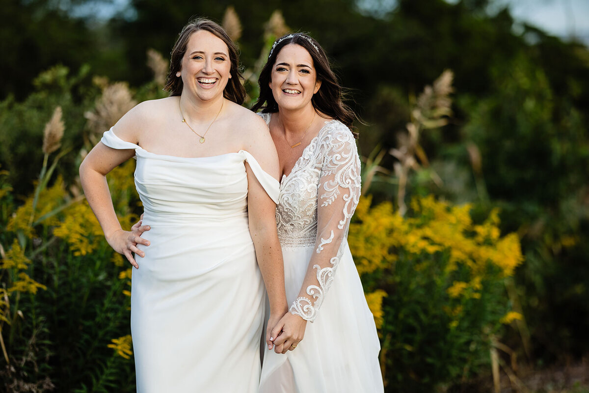 The two brides stand side by side, smiling at the camera, one arm around each other, with a field of tall grass behind them.