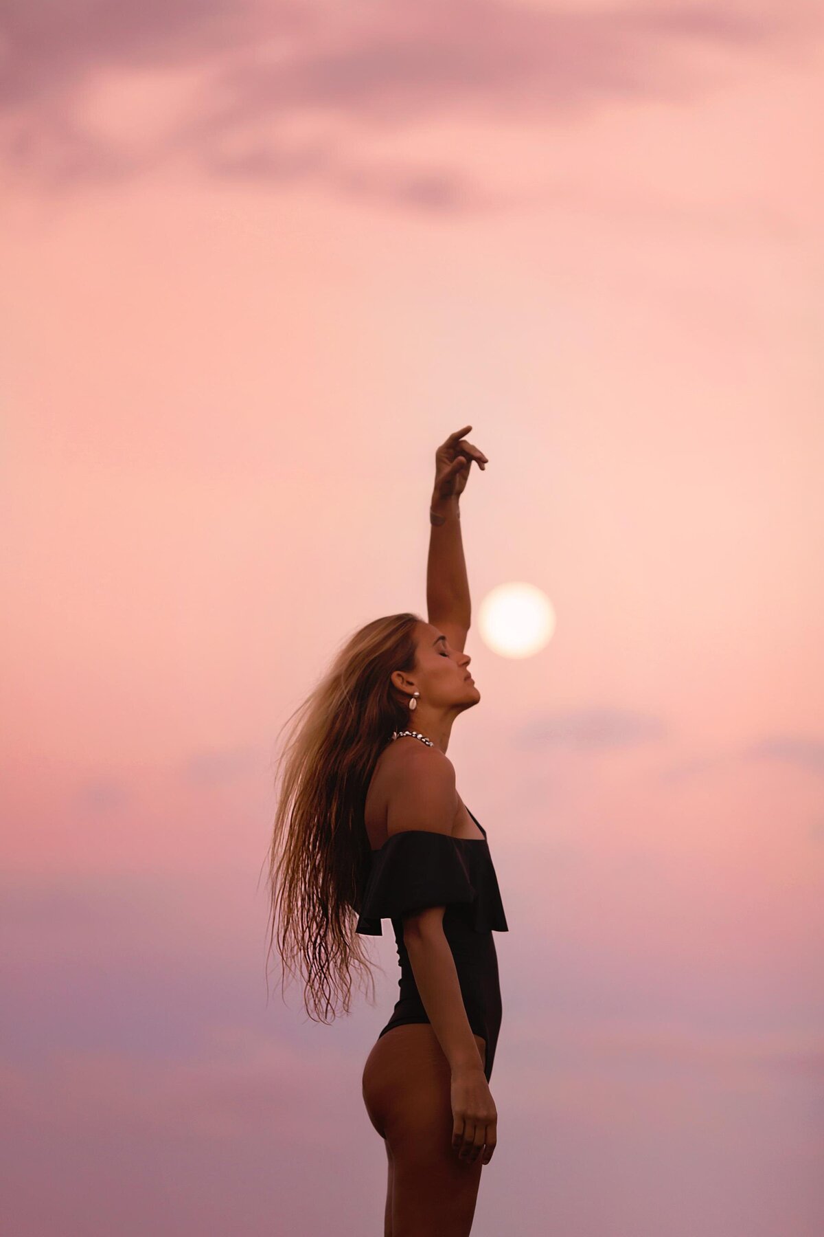 Woman with long hair stands in a black bathing suit with one hand in the air and her face touching the moon at sunset