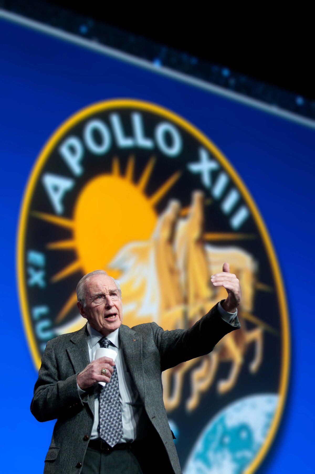 James Lovell speaking about overcoming adversity as he gestuyres with one arm in the air in front of him to simulate Apollo 13