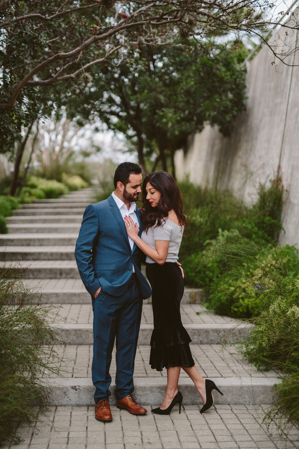 Woman with her hand on fiancé's chest over his heart and standing on steps at the San Antonio Botanical Garden.