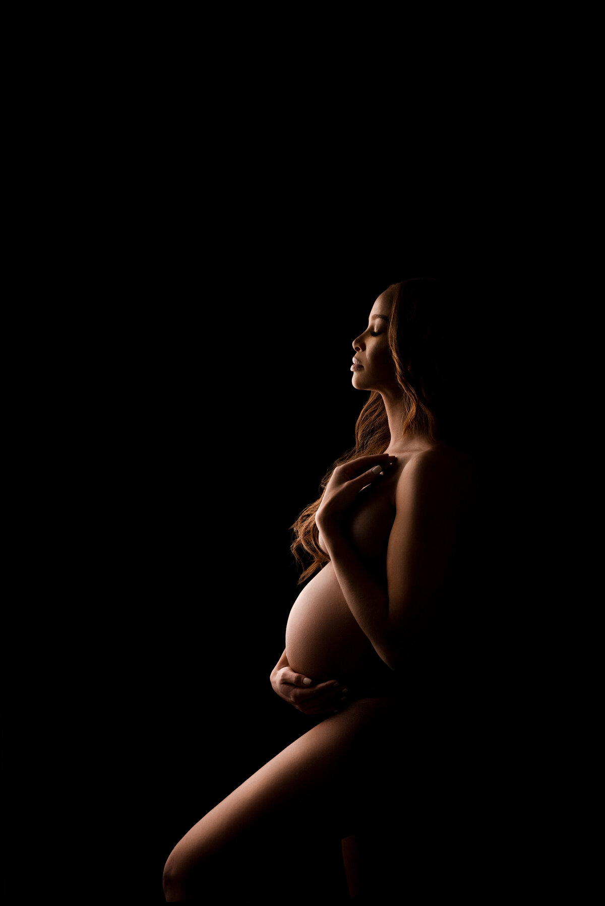 In an intimate fine art maternity photoshoot expertly captured by Katie Marshall, recognized as the best maternity photographer in New Jersey, a striking image unfolds. Here, a woman stands in elegant side profile to the camera, her front knee gracefully bent, and one hand cradling her bump while her forehand gently touches her shoulder. With closed eyes, she turns her face toward the luminous light source, casting captivating highlights and deep shadows that infuse the image with profound depth and dimension.
