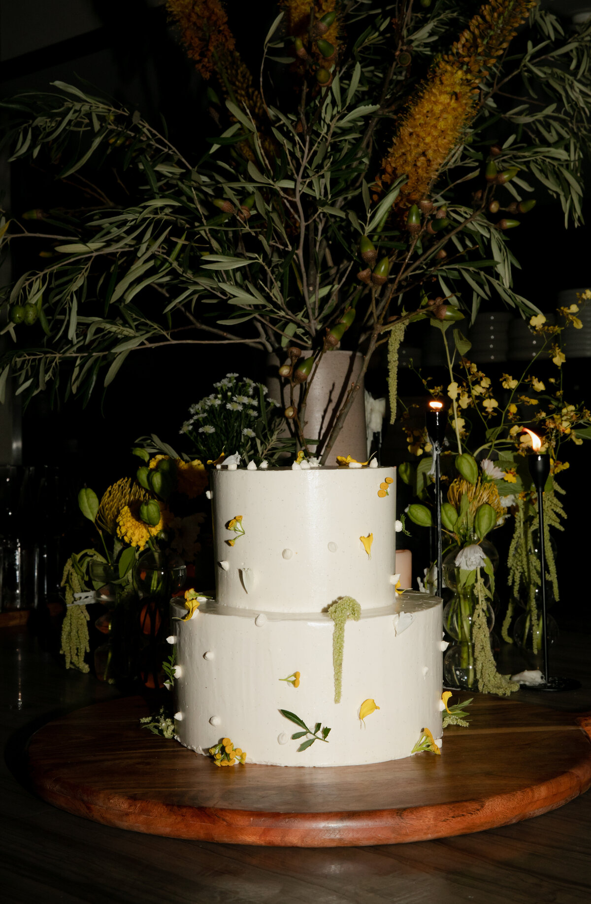 Two tier white wedding cake decorated with yellow florals and lush greenery sit atop a wooden table at Elske Restaurant in Chicago.