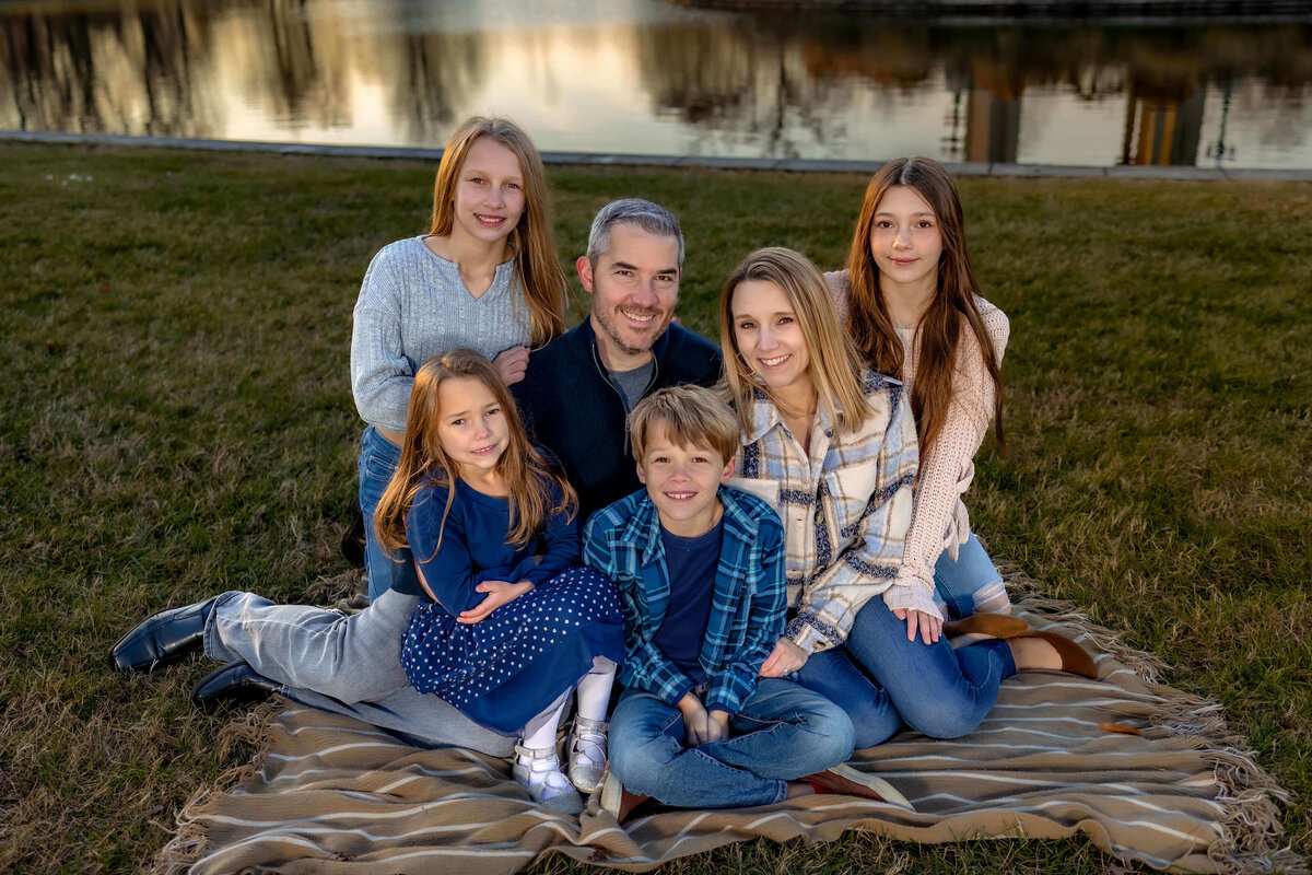Family of 6 sitting on a blanket in a park by a pond at sunset, everyone is looking at the camera