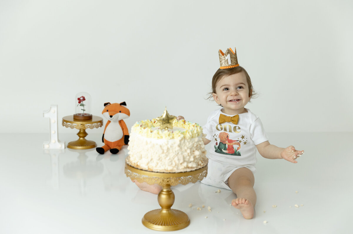 A young toddler birthday boy sits in a studio in a tiny crown and gold bowtie happily playing with a cake