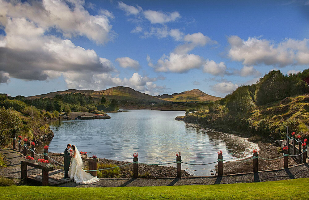 bride wearing a layered tulle, princess style wedding dress kissing her husband wearing a grey tailcoat, while standing on a wooden bridge overlooking Golden Cove at the Sneem Hotel in Kerry