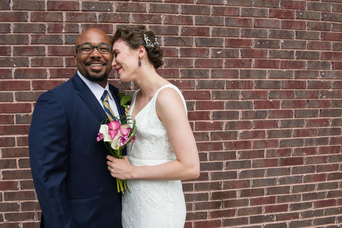Black groom and bride smile in front of brick wall.