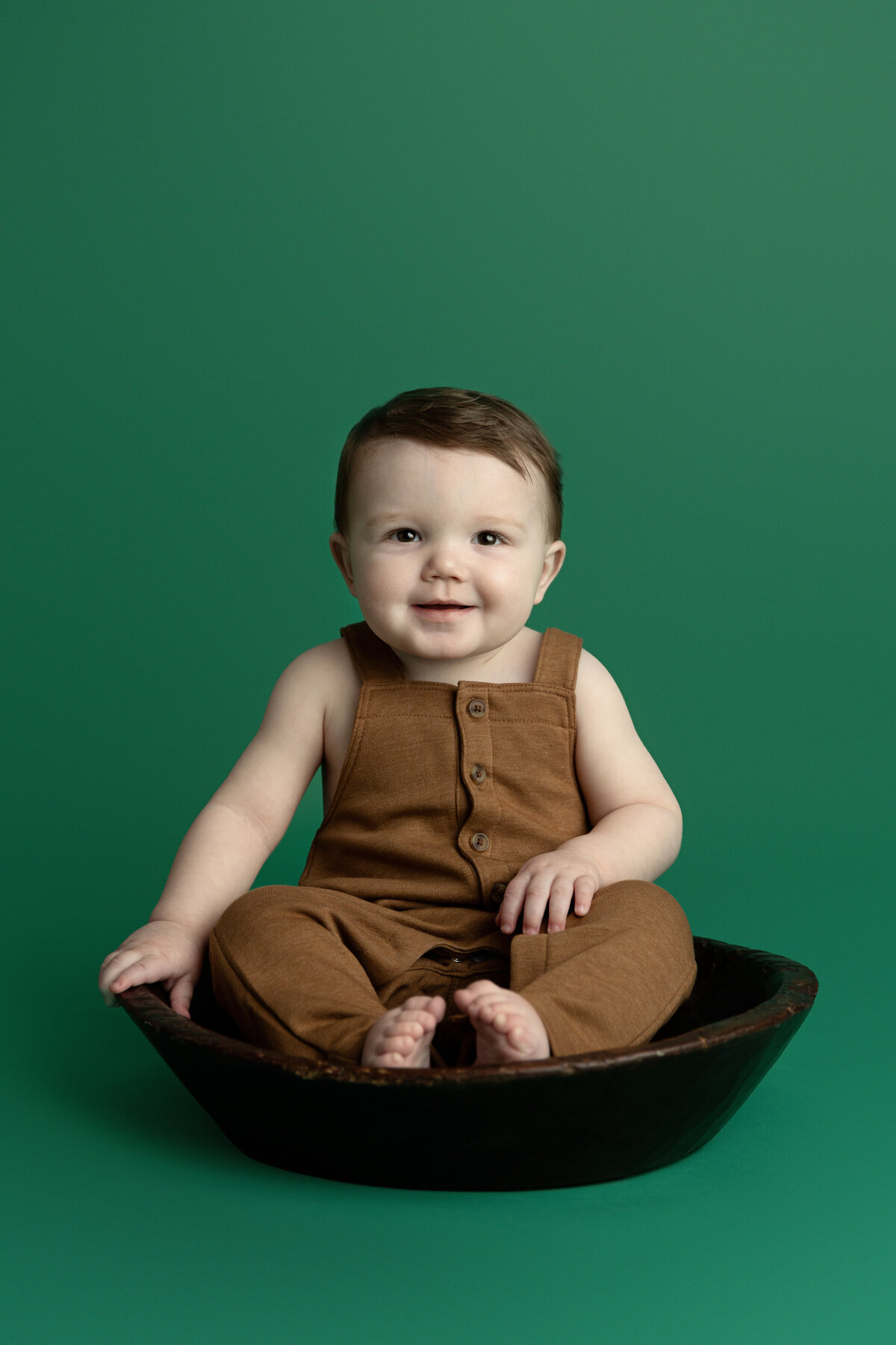A toddler boy in brown overalls sits in a wooden bowl in a green photo studio