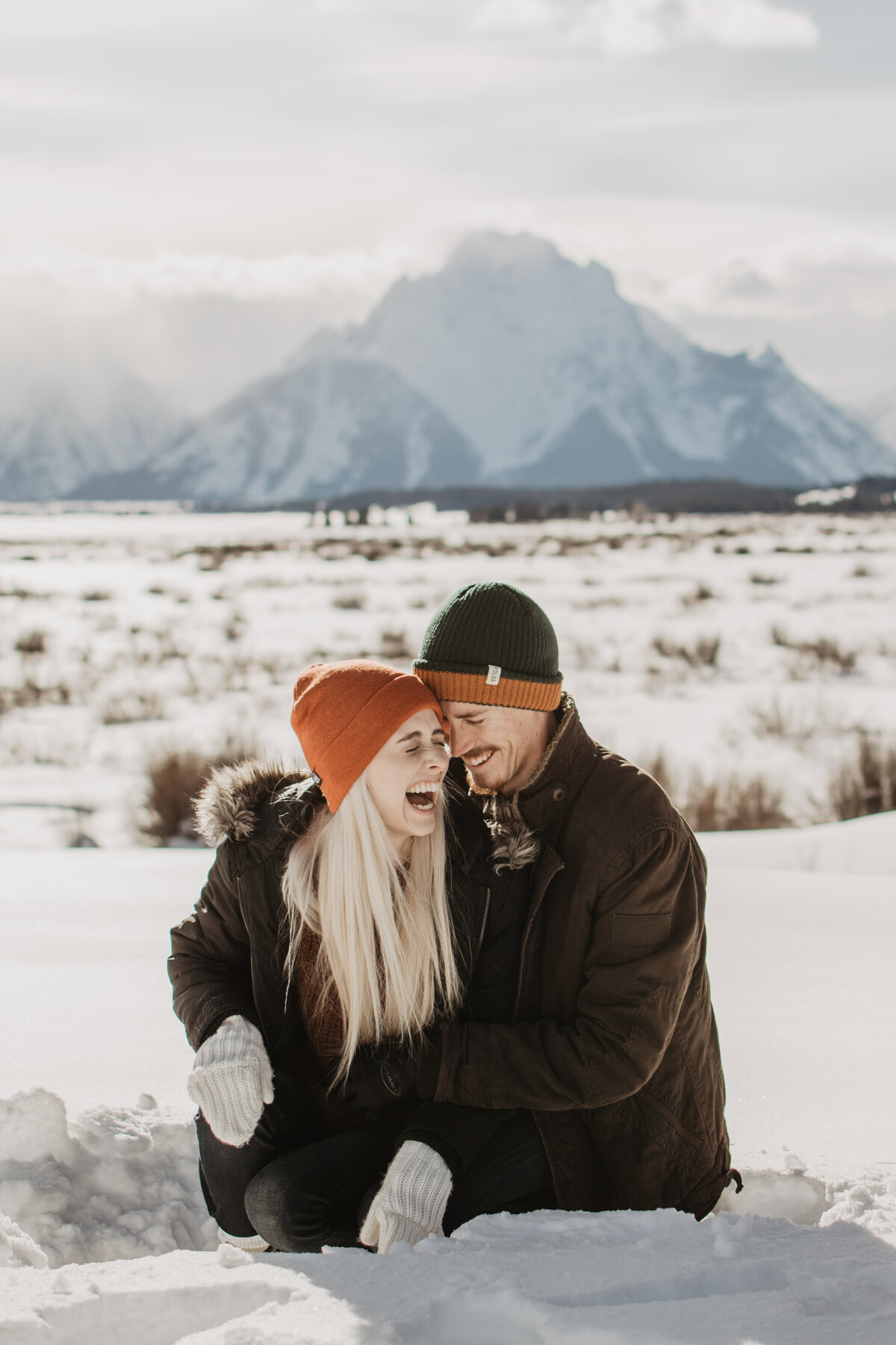 jackson hole photographers captures engagement picture with blonde woman laughing with her fiance as they sit in the snow and tell jokes during their engagement session in winter in Jackson Hole Wyoming