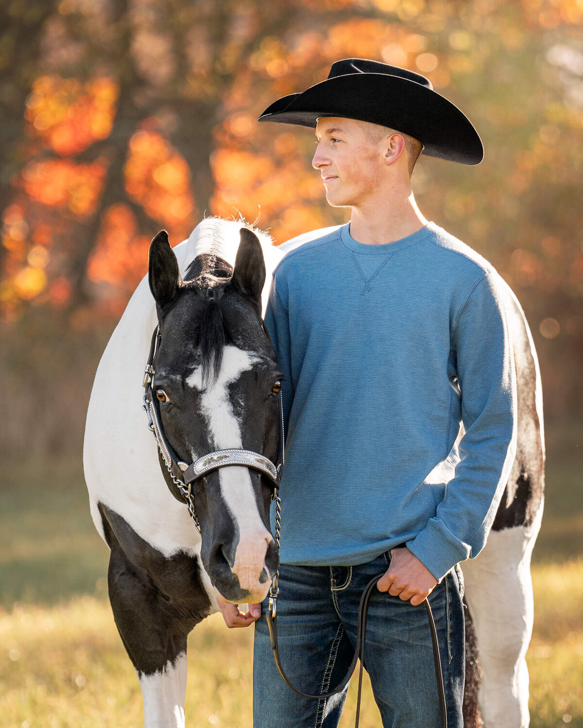 male equestrian wearing a long sleeve blue shirt, jeans, and a black cowboy had standing proudly next to his black and white tobiano Paint horse gelding looking away from the camera with beautiful colored fall leaves behind them.