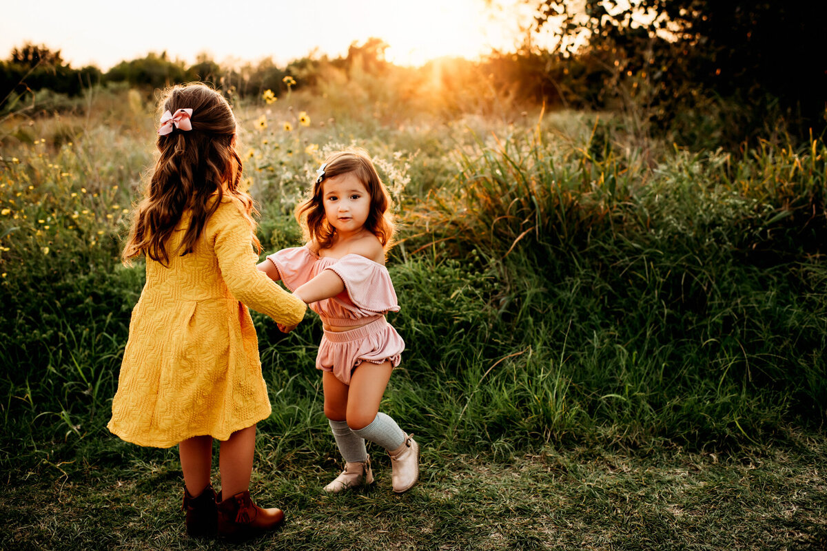 Family Photographer, two young girls dance together near tall green grass