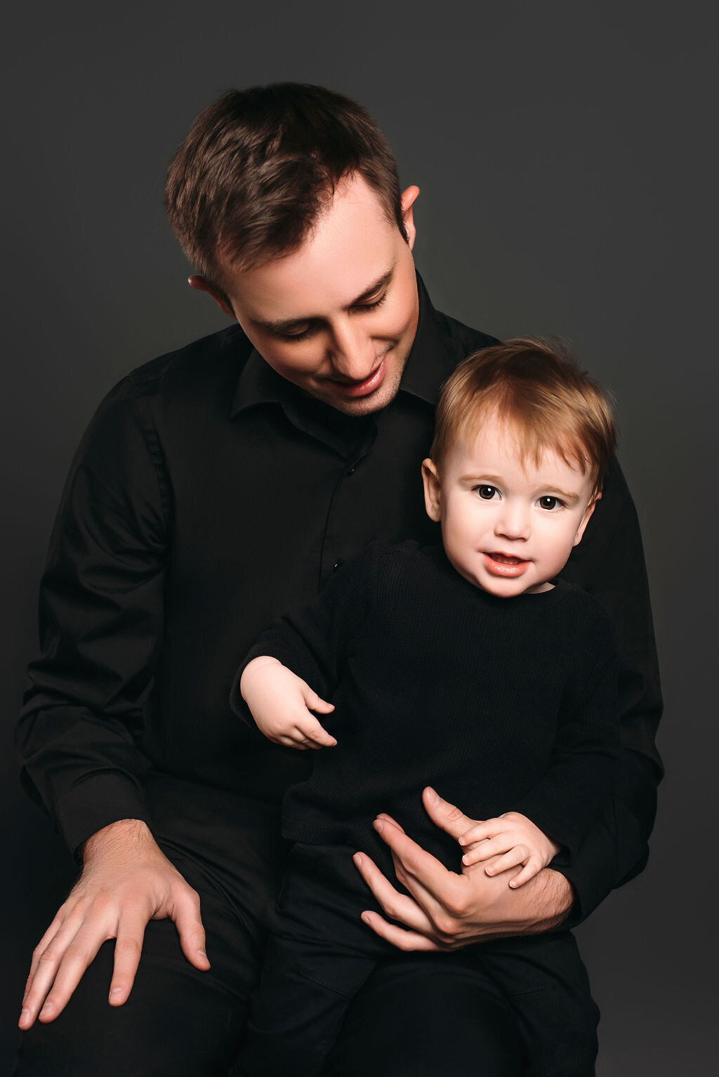 A father in black dress clothes stands in front of a gray-toned backdrop holding his young son in a black outfit, captured by a photographer in Bemidji, Minnesota.