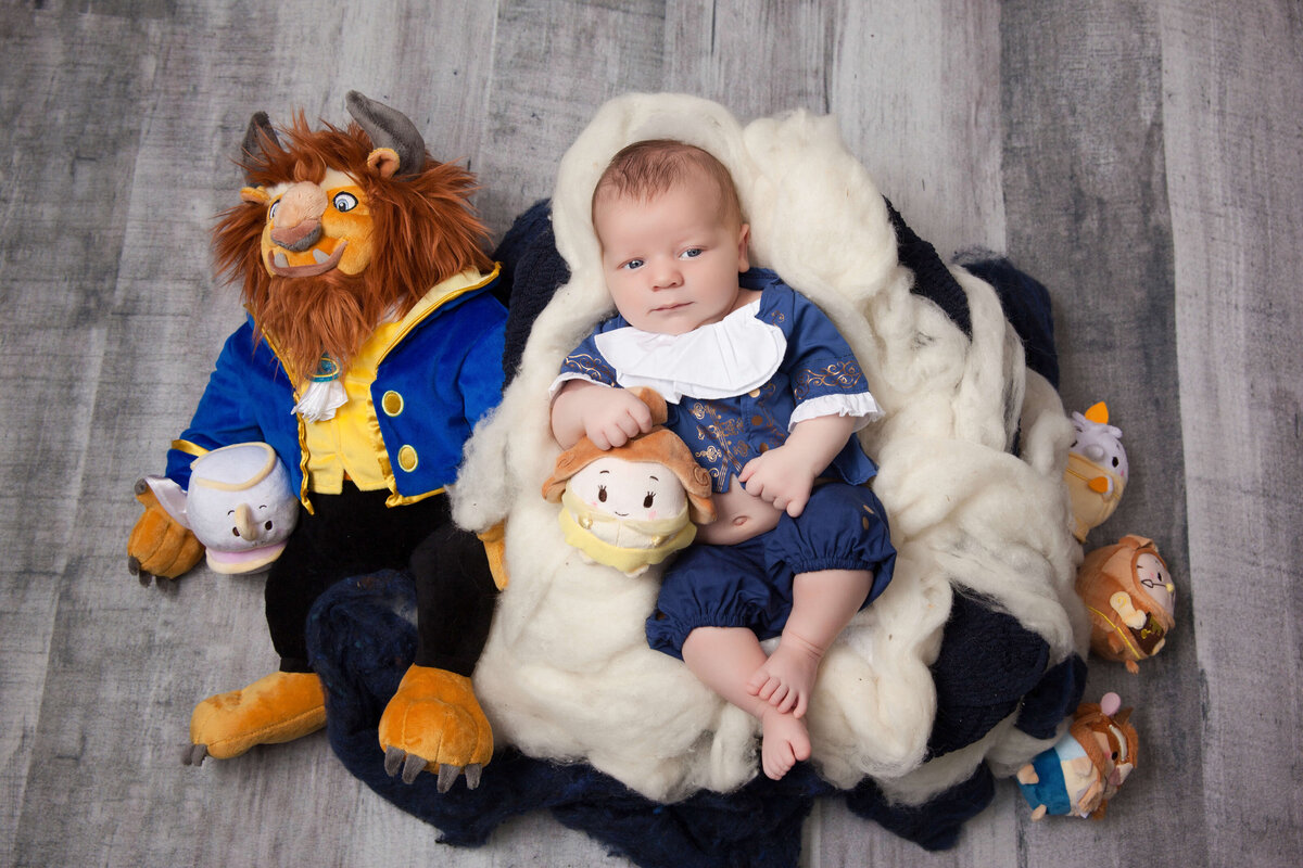 A baby lies on a fluffy, cream-colored blanket, surrounded by plush toys. The largest toy is a beast with horns and a blue coat. The baby wears a blue outfit and holds a smaller plush character. Captured beautifully by a Jacksonville newborn photographer, the background is a gray wooden floor.