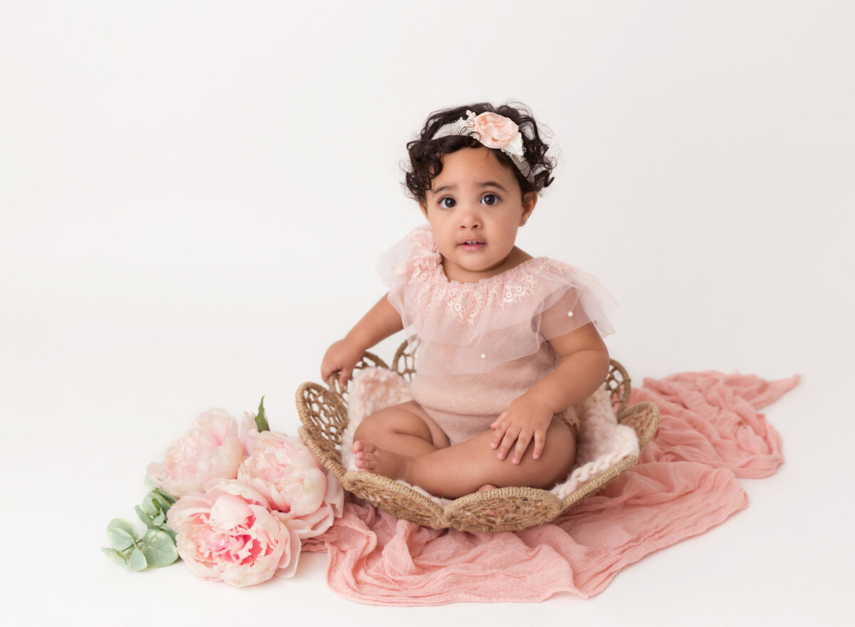 Baby girl sits in rattan basket for baby photoshoot. Baby is looking at the camera wearing a blush onesie with organza detail.