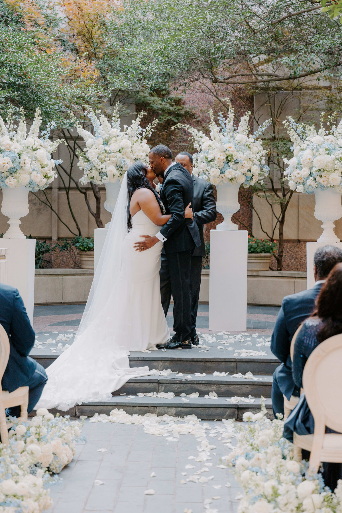 A bride and groom share a kiss on an outdoor altar adorned with white and blue flowers. Guests watch from decorated chairs, and petals line the aisle.