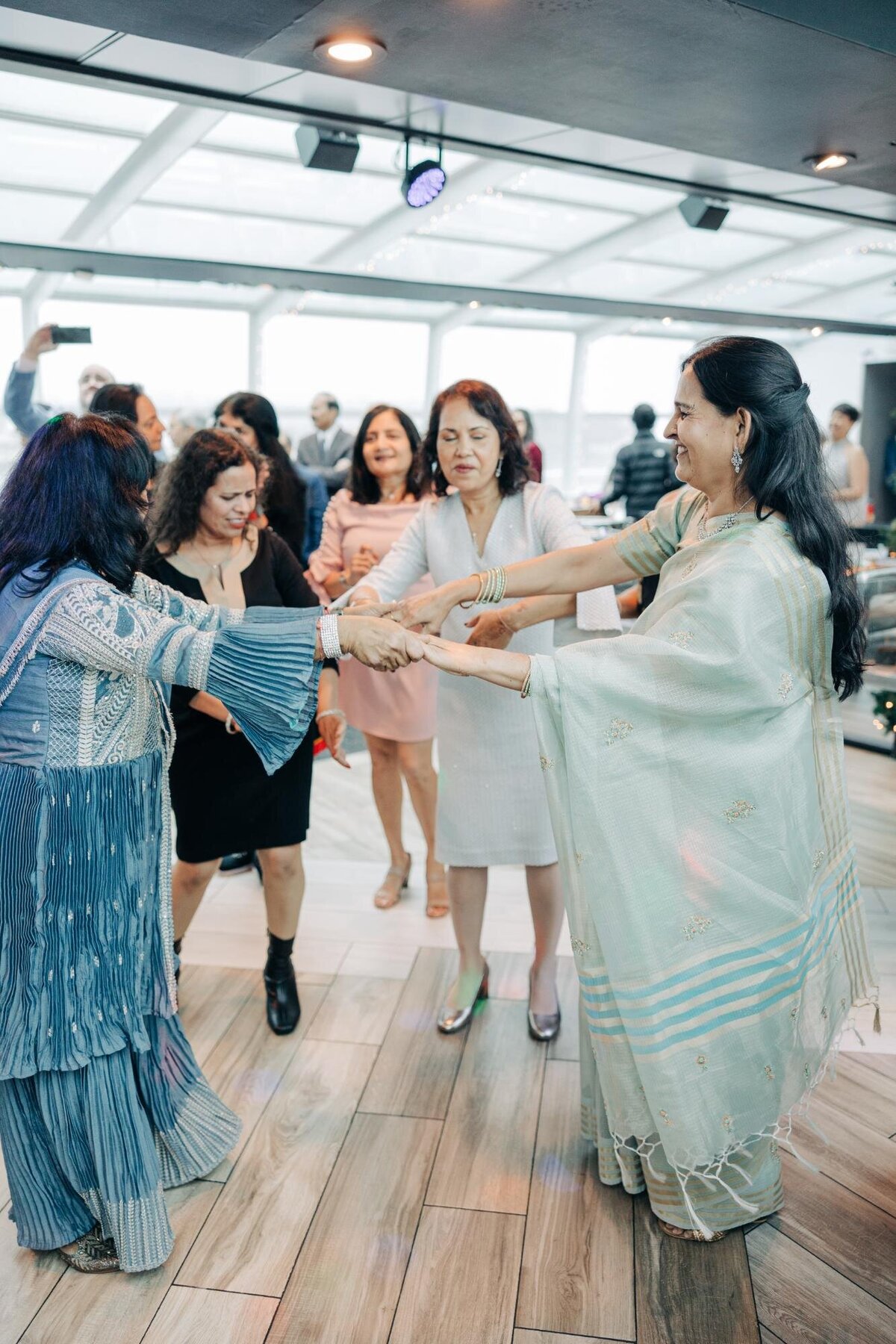 Women in traditional and casual attire dancing in a circle at an indoor event, smiling and holding hands.