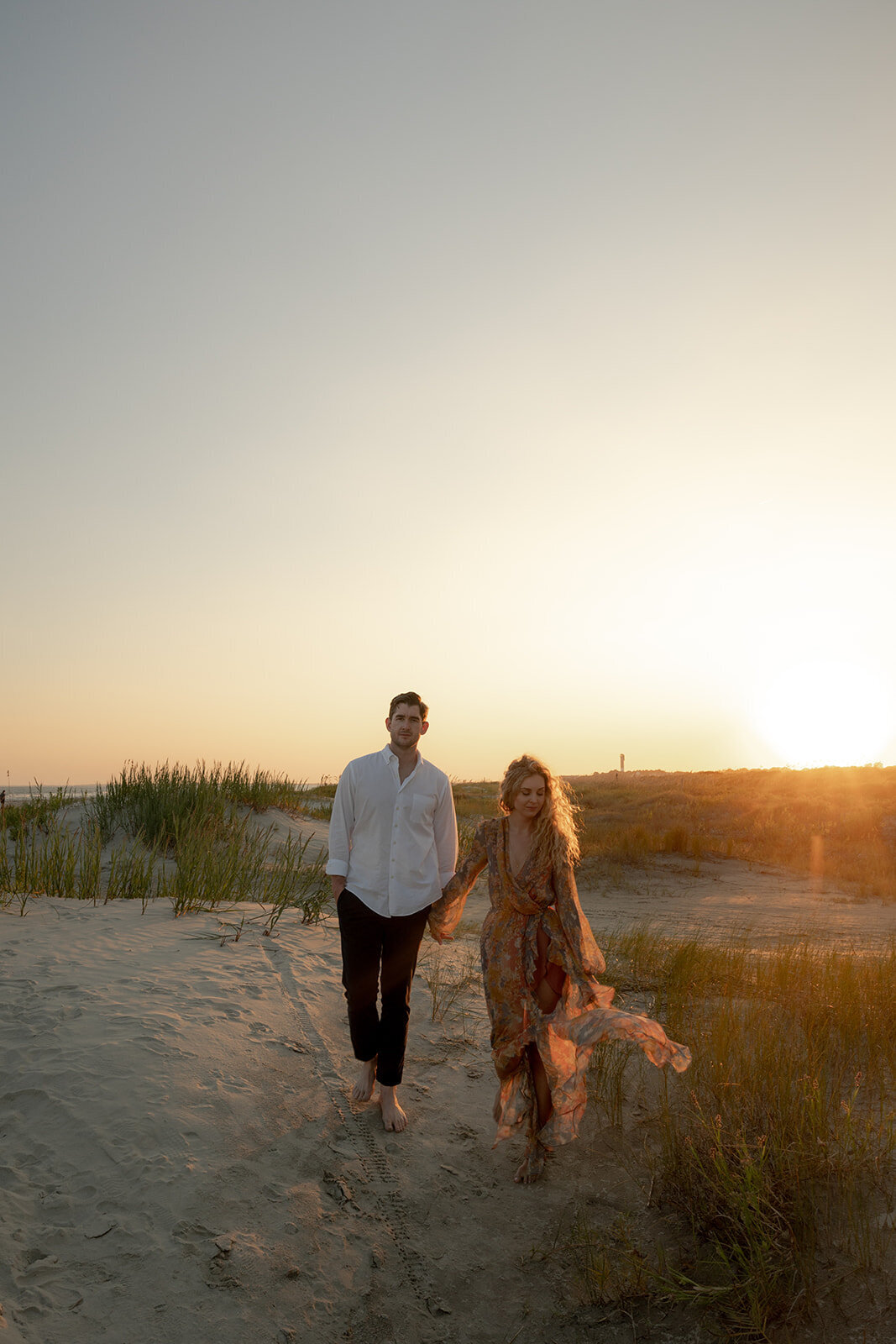 Couple is walking in sand dunes during Charleston beach engagement session. Sun is setting. Woman is wearing flowy dress perfectly blending in with warm color created by sunset.