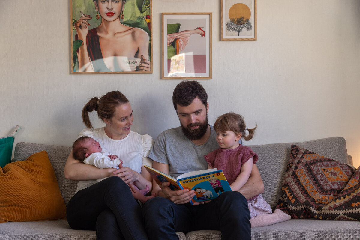 Familie på fire sitter i sofaen; mor med baby på armen og far med storesøster i armkroken, de leser bok sammen.