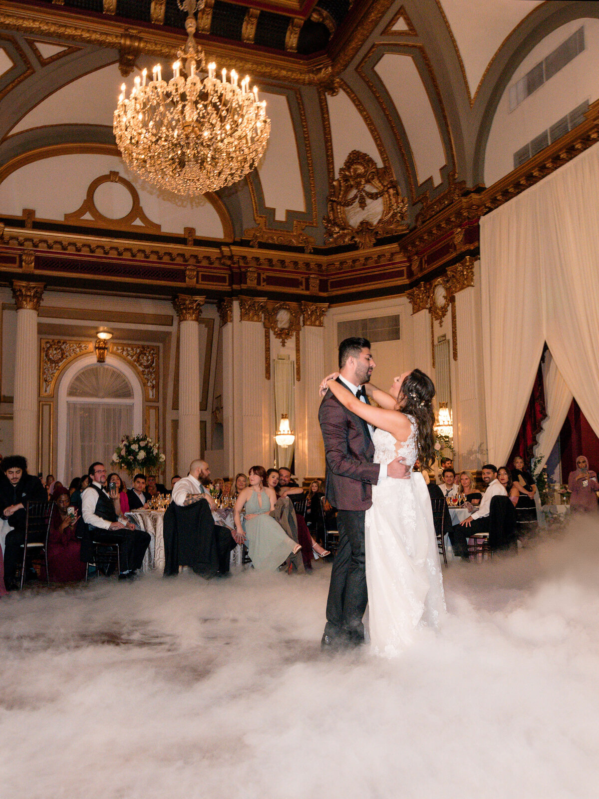 A couple shares their first dance at a wedding in an opulent ballroom with a large chandelier. Guests seated around the room watch as the floor is covered in fog, adding to the romantic atmosphere.