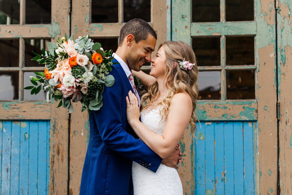 Wedding-couple-cuddle-on-wedding-day-in-front-of-blue-doors-with-colorful-flower-bouquet-at-Twin-Willow-Gardens-wedding-venue-in-Snohomish-WA-photo-by-Joanna-Monger-Photography