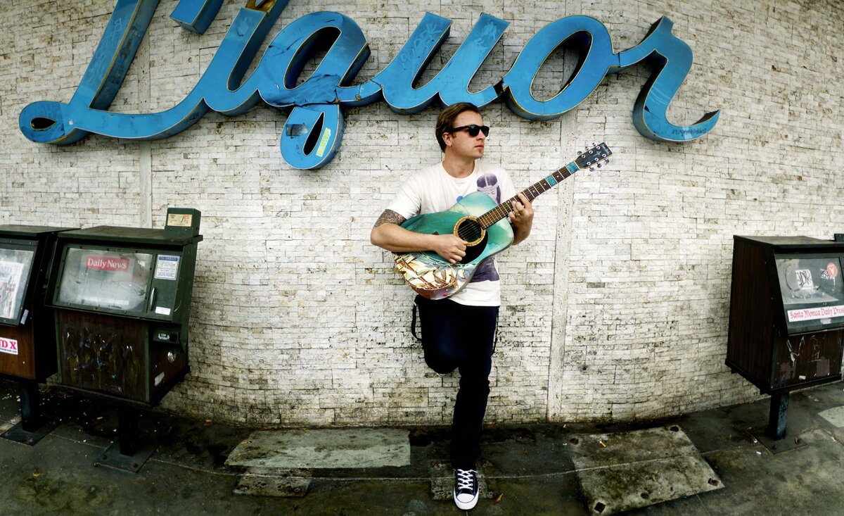 Male musician photo Daniel Wesley wearing white shirt playing blue guitar leaning against white stone wall blue liquor sign behind with newspaper boxes