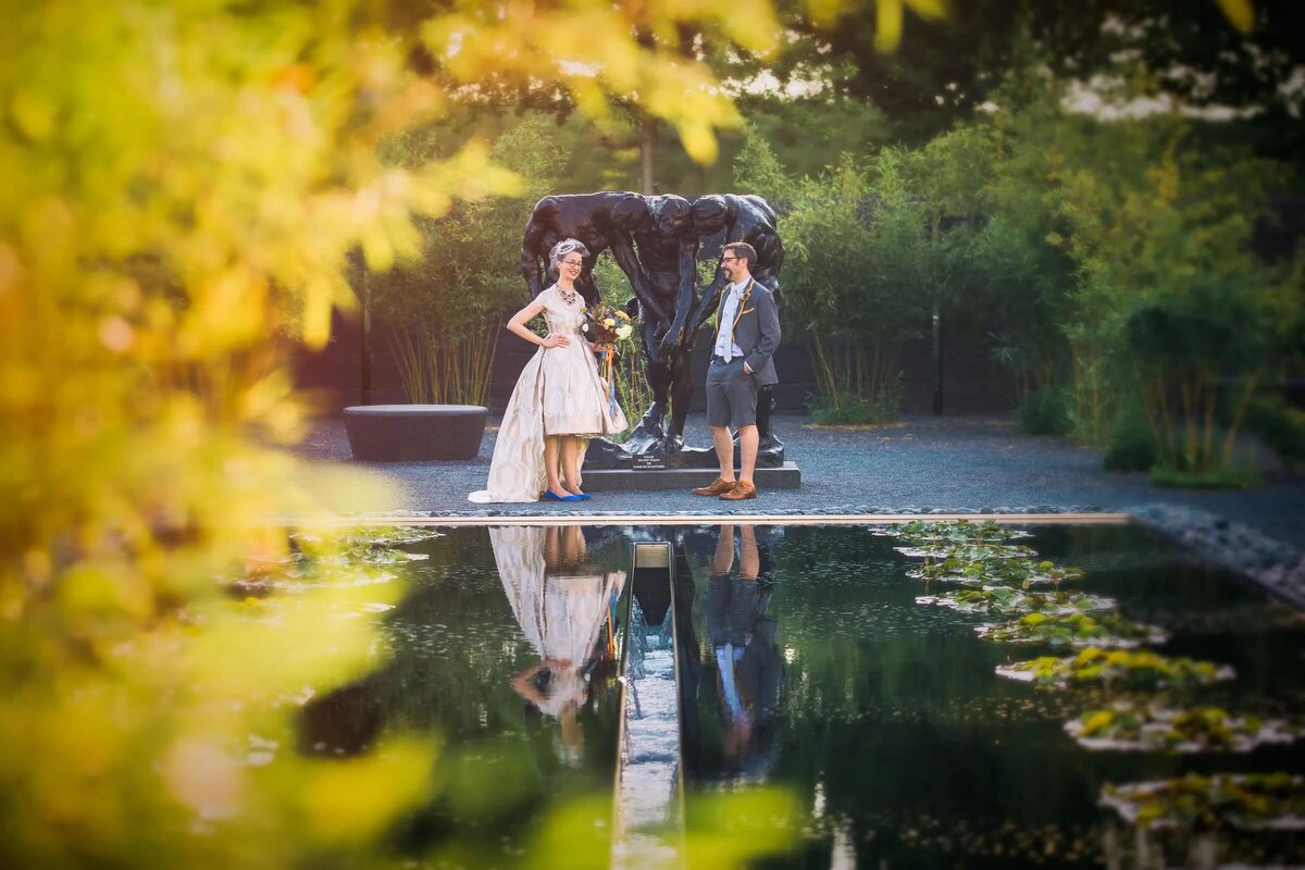 A couple exchanges vows by a reflective pond in a lush garden, with soft light filtering through the trees.
