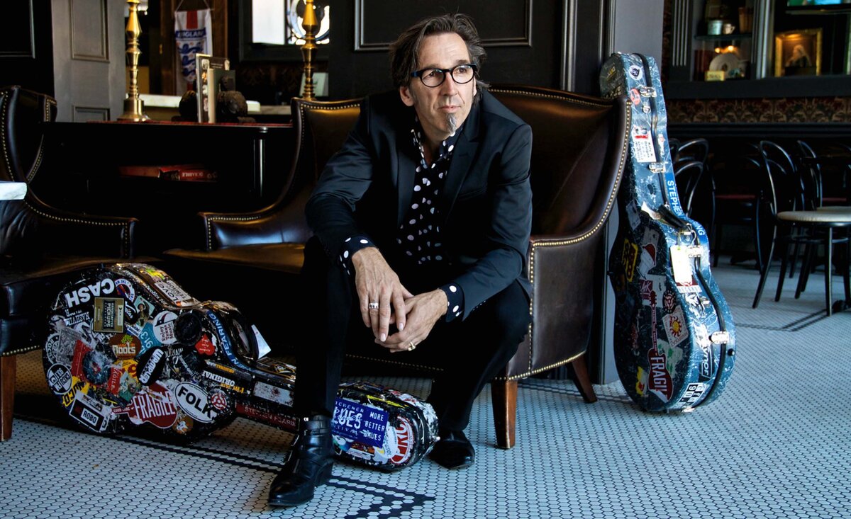 Male musician portrait Stephen Fearing wearing black suit polka shirt sitting between sticker covered guitar cases