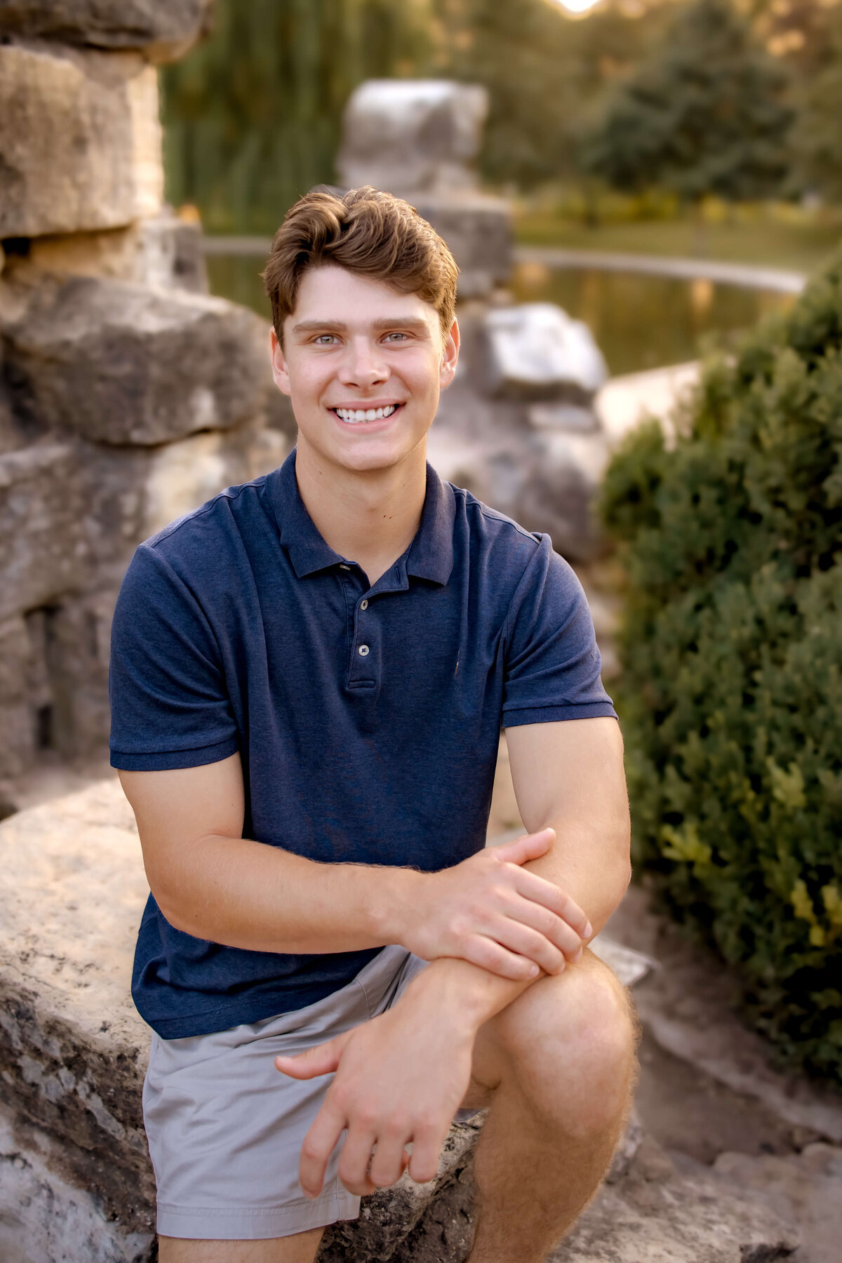 A handsome young man is posed on a stone structure in a park for his senior portraits.