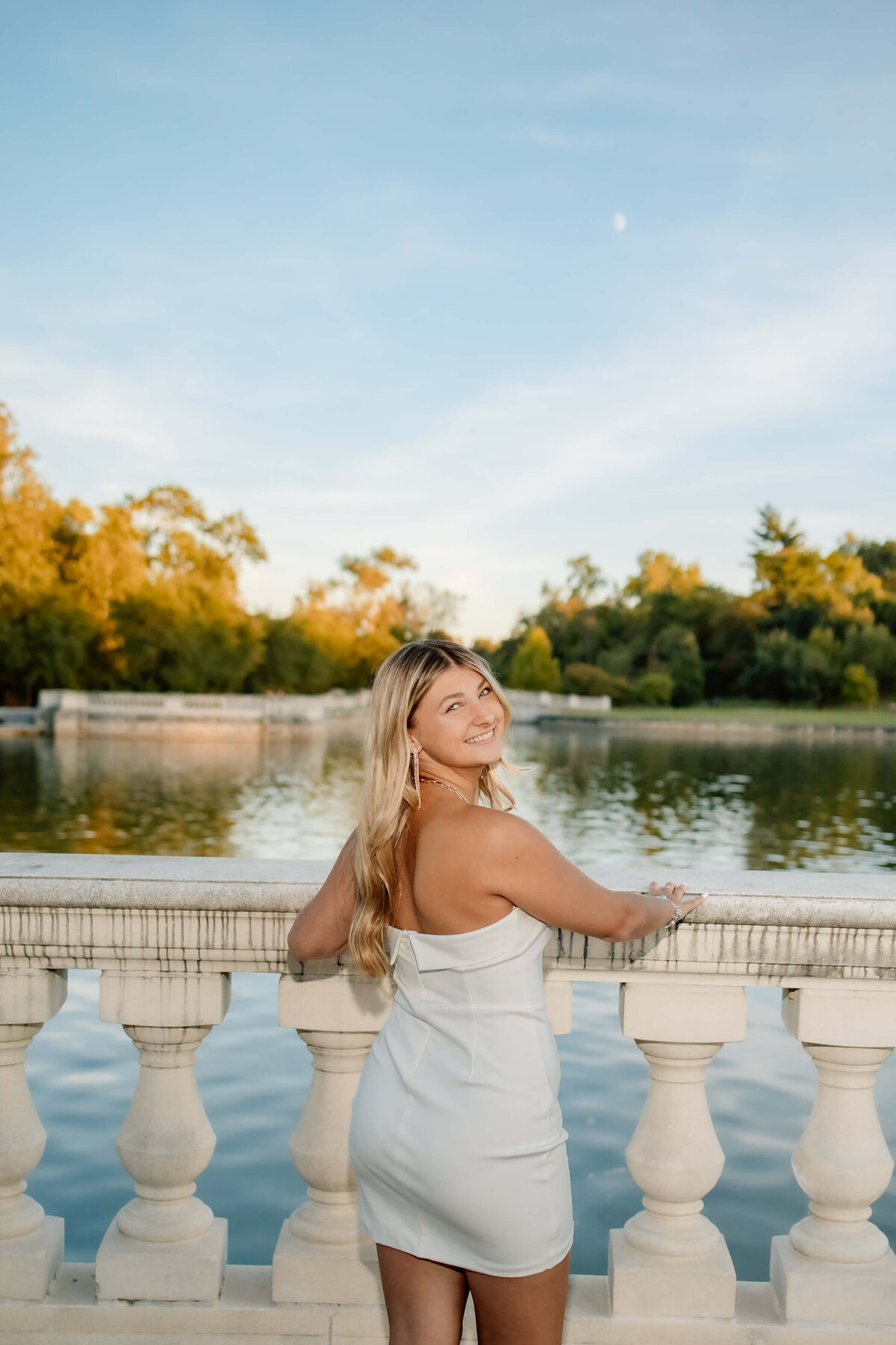 a blonde haired girl in a tight white dress  standing by a cement railing  next to a lake at sunset