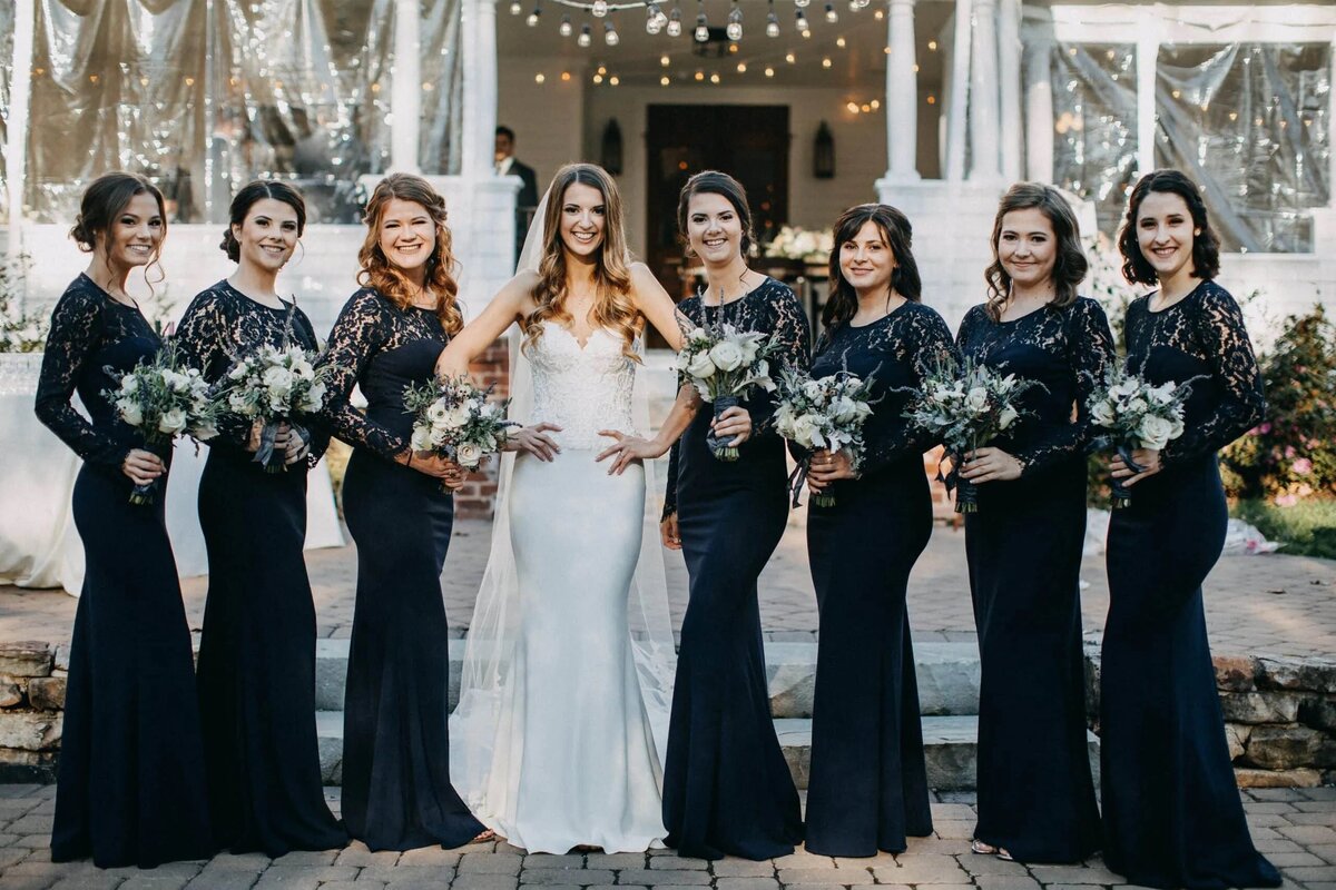 A bride stands with her bridesmaids, who are dressed in elegant black dresses, all holding bouquets and smiling at the camera.
