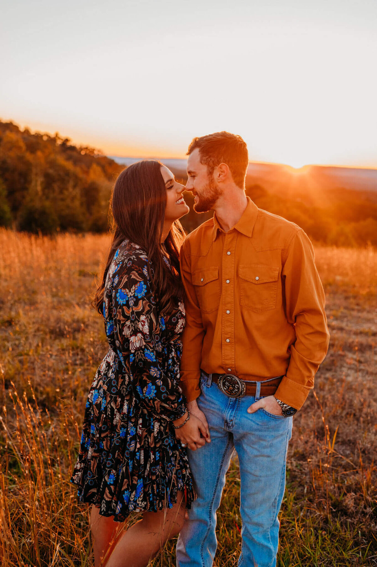 photo of of man in an orange button-down shirt and a woman holding his hand and gicing eskimo kisses in a field