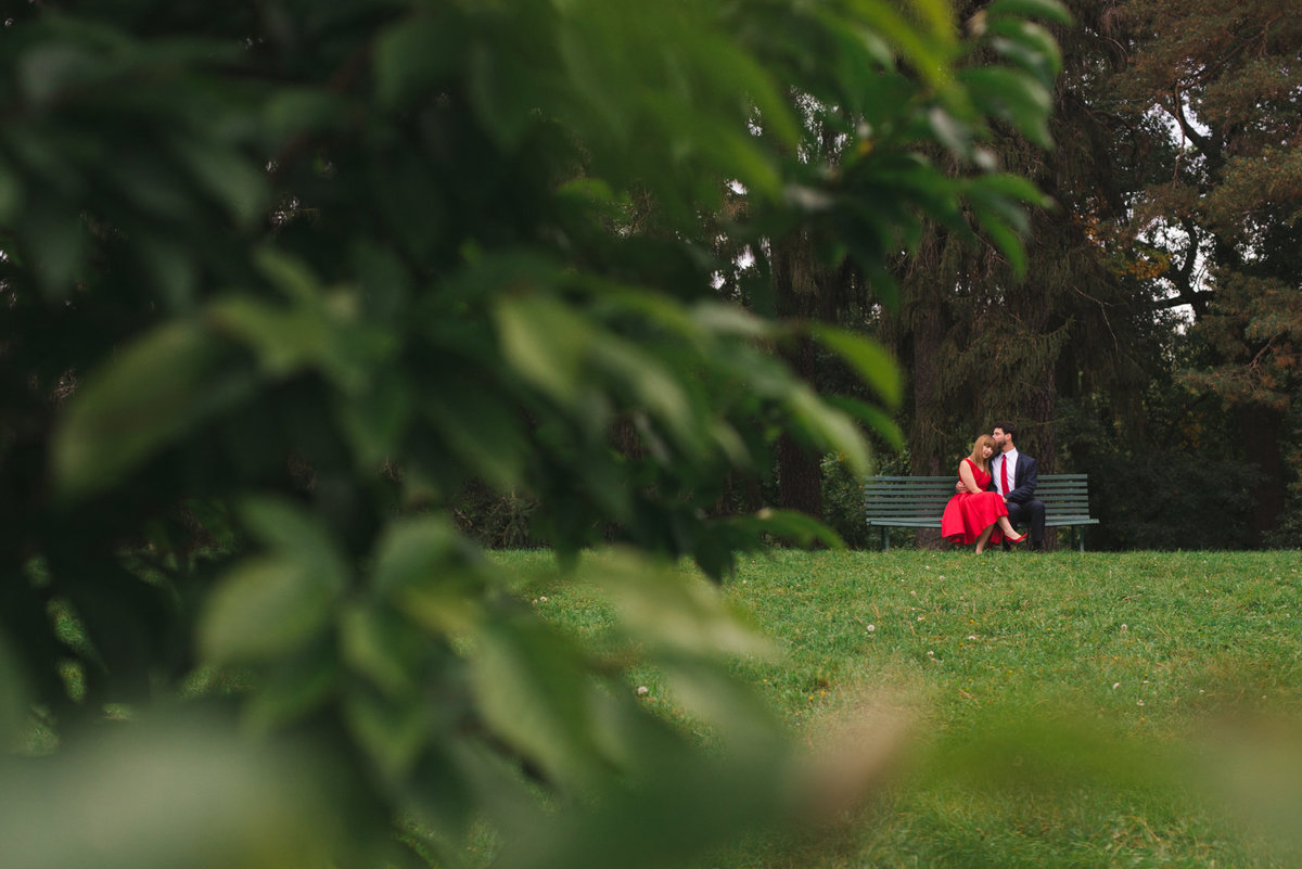 Girl wearing red dress sitting on a bench with her fiancee among the trees