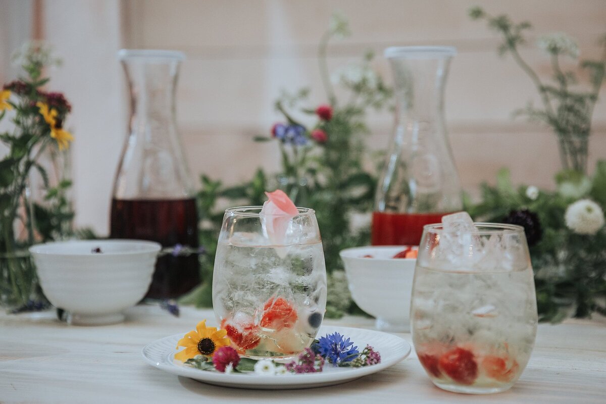 two clear highball cocktails with flowers encased in ice cubes. A white plate with a rainbow of flowers sitting on a white farm table. Carafes of wine sitting in the background.
