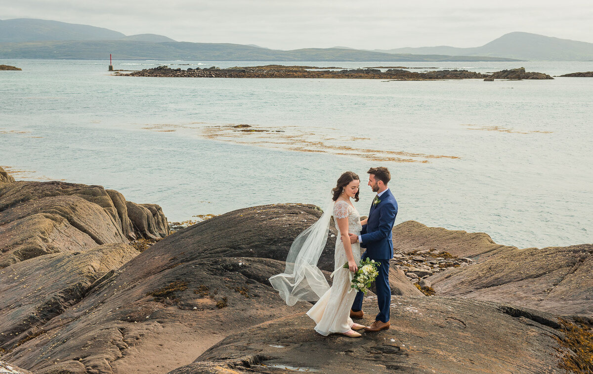 Bride with black hair, holding a white bouquet and wearing a vintage, sequenced dress with groom in blue suit standing on rocks overlooking the Atlantic ocean