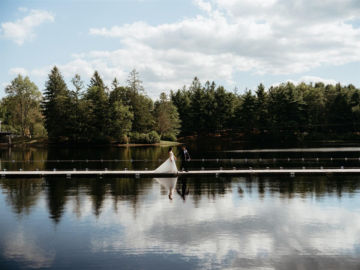 Dramatic wide shot portrait of bride and groom facing each other on long dock in the middle of a calm lake with trees in background, blue skies, and clouds reflecting in water.