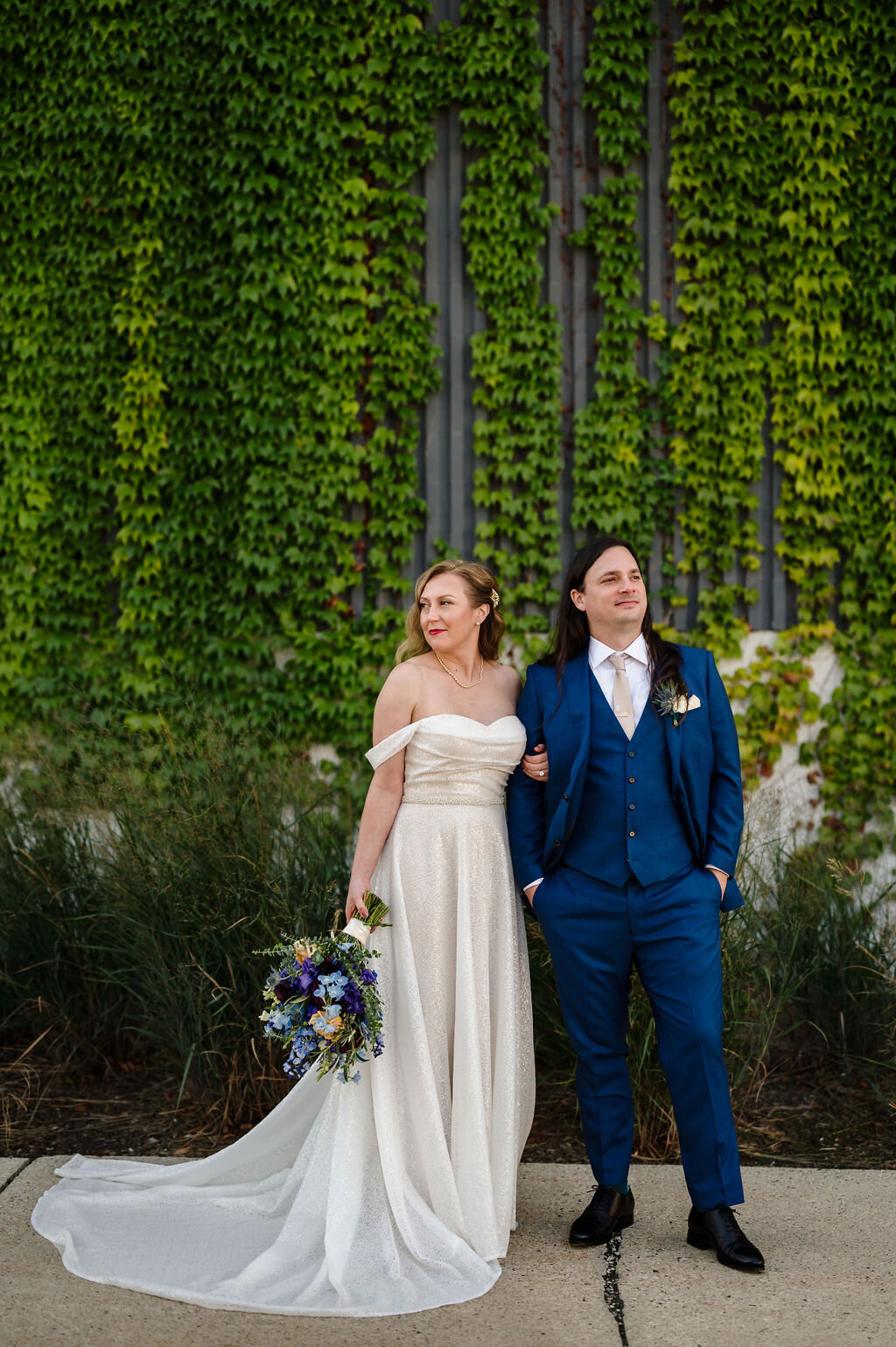 Bride and groom stand in front of  ivy wall