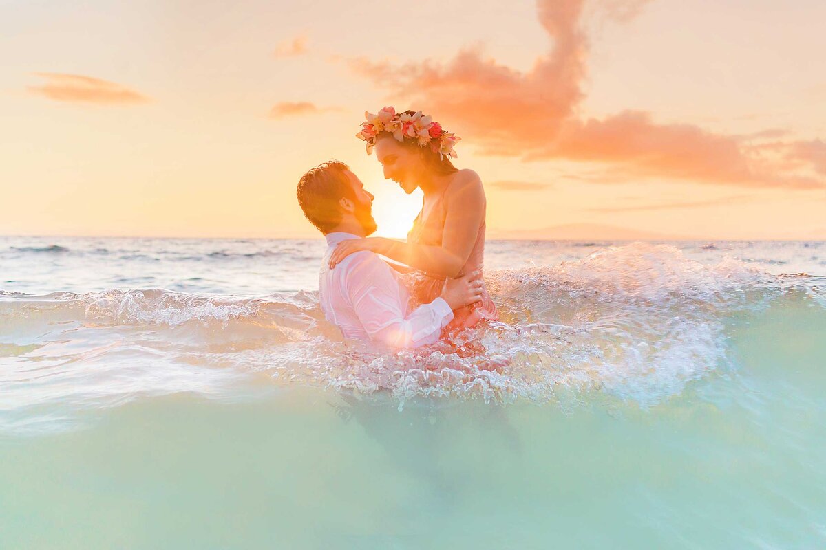 Man lifts his wife over a wave while in the ocean at sunset, being photographed by Love + Water for their honeymoon portraits in Hawaii