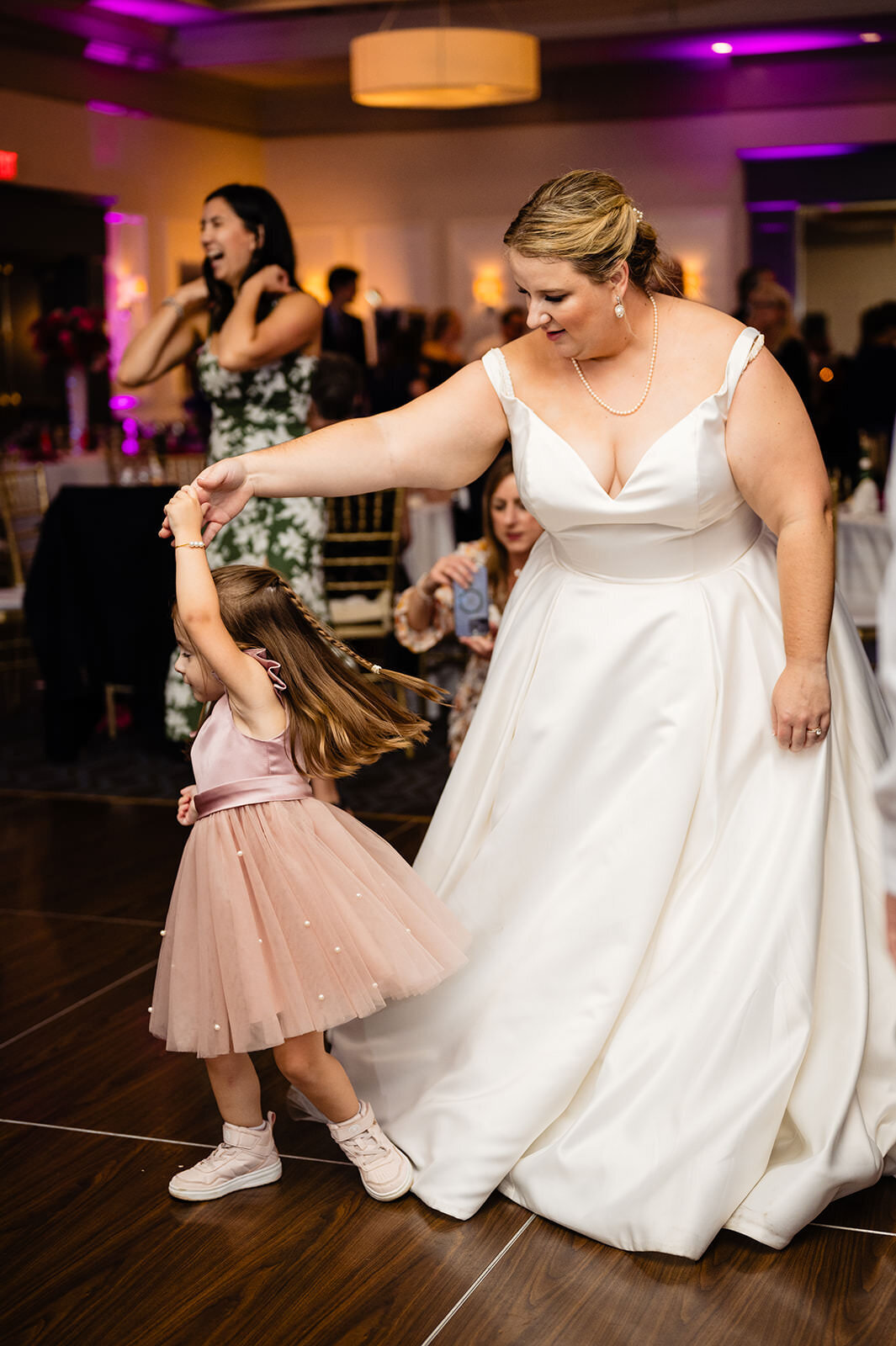A bride joyfully dances with a young girl in a pink dress, swinging her around the dance floor.