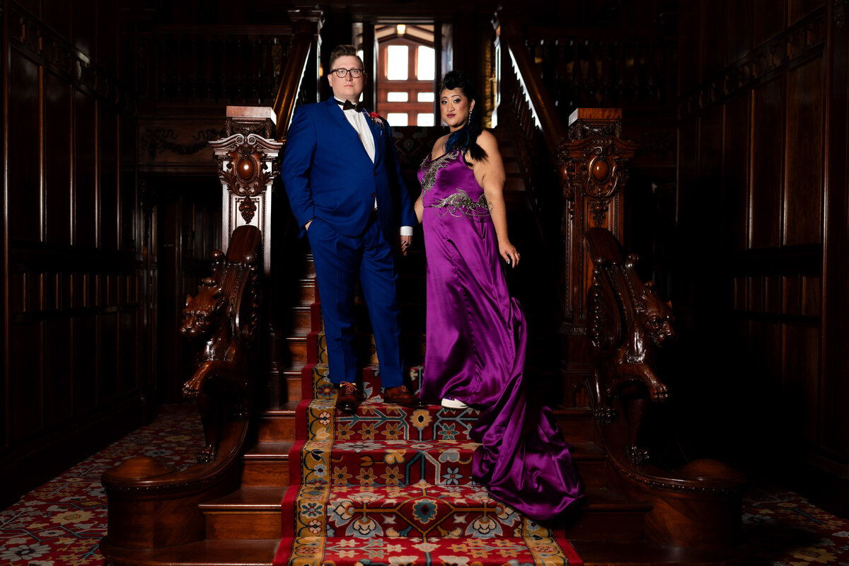 Bride in purple dress and groom in blue suit stand on stairs.